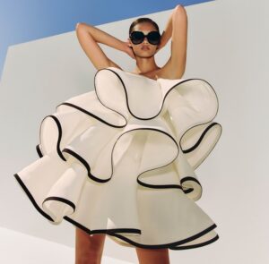 A woman wearing dark sunglasses poses with both arms raised behind her head against a white wall and blue sky background. She's wearing a dramatic white dress or top with voluminous, sculptural ruffles and black piping trim that creates an avant-garde, architectural silhouette.