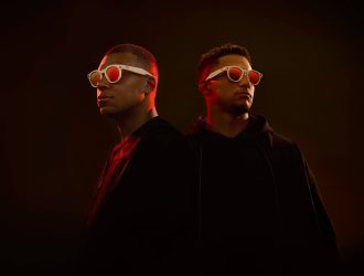 Kylian Mbappé et Patrick Mahomes se tiennent côte à côte sur un fond sombre avec un éclairage rouge dramatique. Mbappé (à gauche) porte une tenue noire à col ras du cou tandis que Mahomes (à droite) porte un sweat à capuche noir. Les deux athlètes portent des lunettes intelligentes Oakley x Meta à monture blanche et aux verres teintés d'orange. L'éclairage sombre crée un contraste saisissant, soulignant le design futuriste des lunettes Oakley Meta dans cette photo promotionnelle.