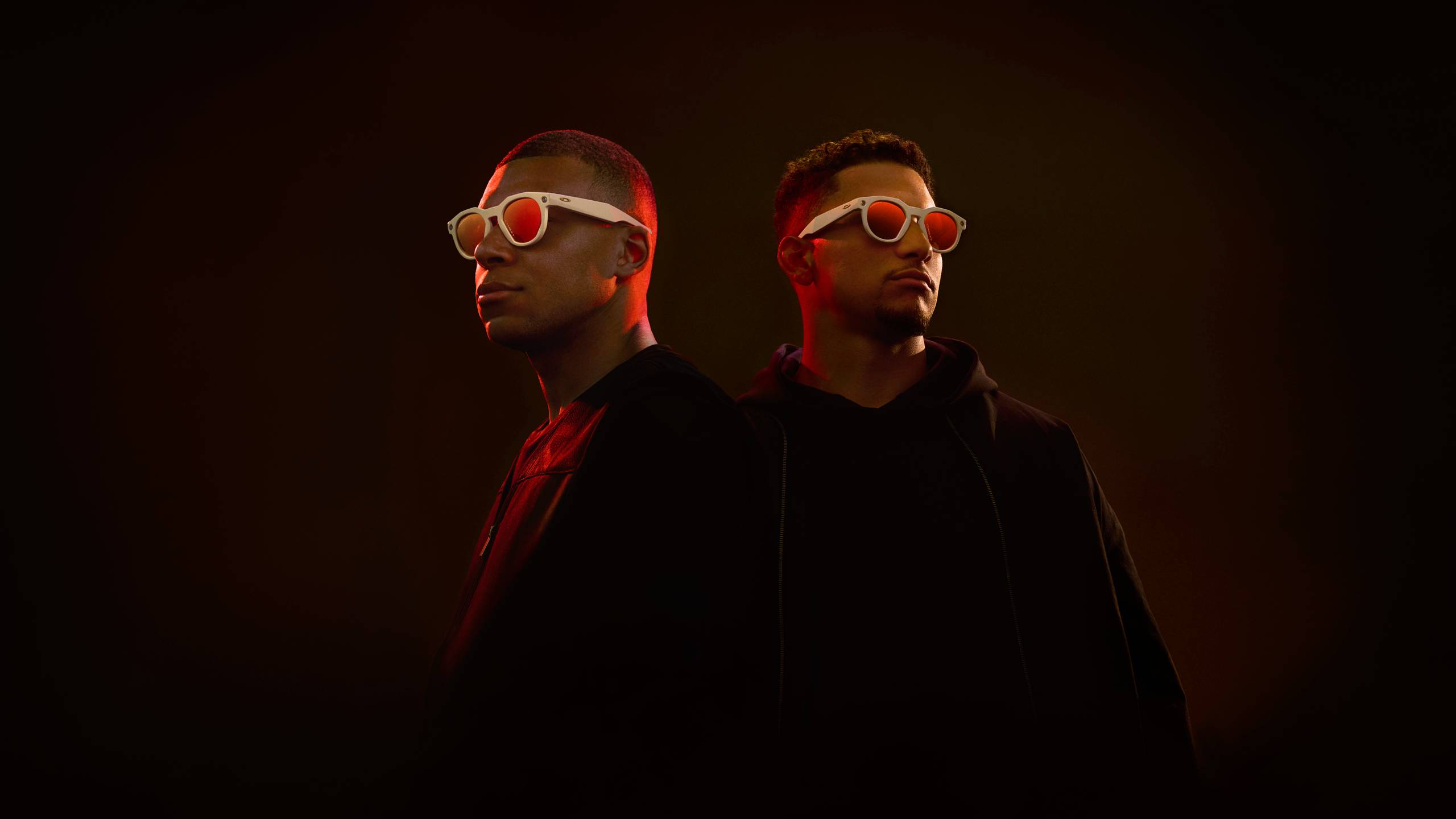 Kylian Mbappé et Patrick Mahomes se tiennent côte à côte sur un fond sombre avec un éclairage rouge dramatique. Mbappé (à gauche) porte une tenue noire à col ras du cou tandis que Mahomes (à droite) porte un sweat à capuche noir. Les deux athlètes portent des lunettes intelligentes Oakley x Meta à monture blanche et aux verres teintés d'orange. L'éclairage sombre crée un contraste saisissant, soulignant le design futuriste des lunettes Oakley Meta dans cette photo promotionnelle.