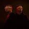 Kylian Mbappé and Patrick Mahomes stand side by side against a dark background with dramatic red lighting. Mbappé (left) wears a black crew neck shirt while Mahomes (right) wears a black hoodie. Both athletes are wearing white-framed Oakley x Meta smart glasses with orange-tinted lenses. The moody lighting creates a striking contrast, emphasizing the futuristic design of the Oakley Meta eyewear in this promotional photograph.