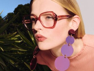 Une femme blonde portant des lunettes à monture rouge surdimensionnée et des boucles d'oreilles circulaires violettes pose sur un fond de palmiers verts et de ciel bleu. Elle porte un pull en maille rose et affiche une expression subtile et confiante en regardant vers la camera