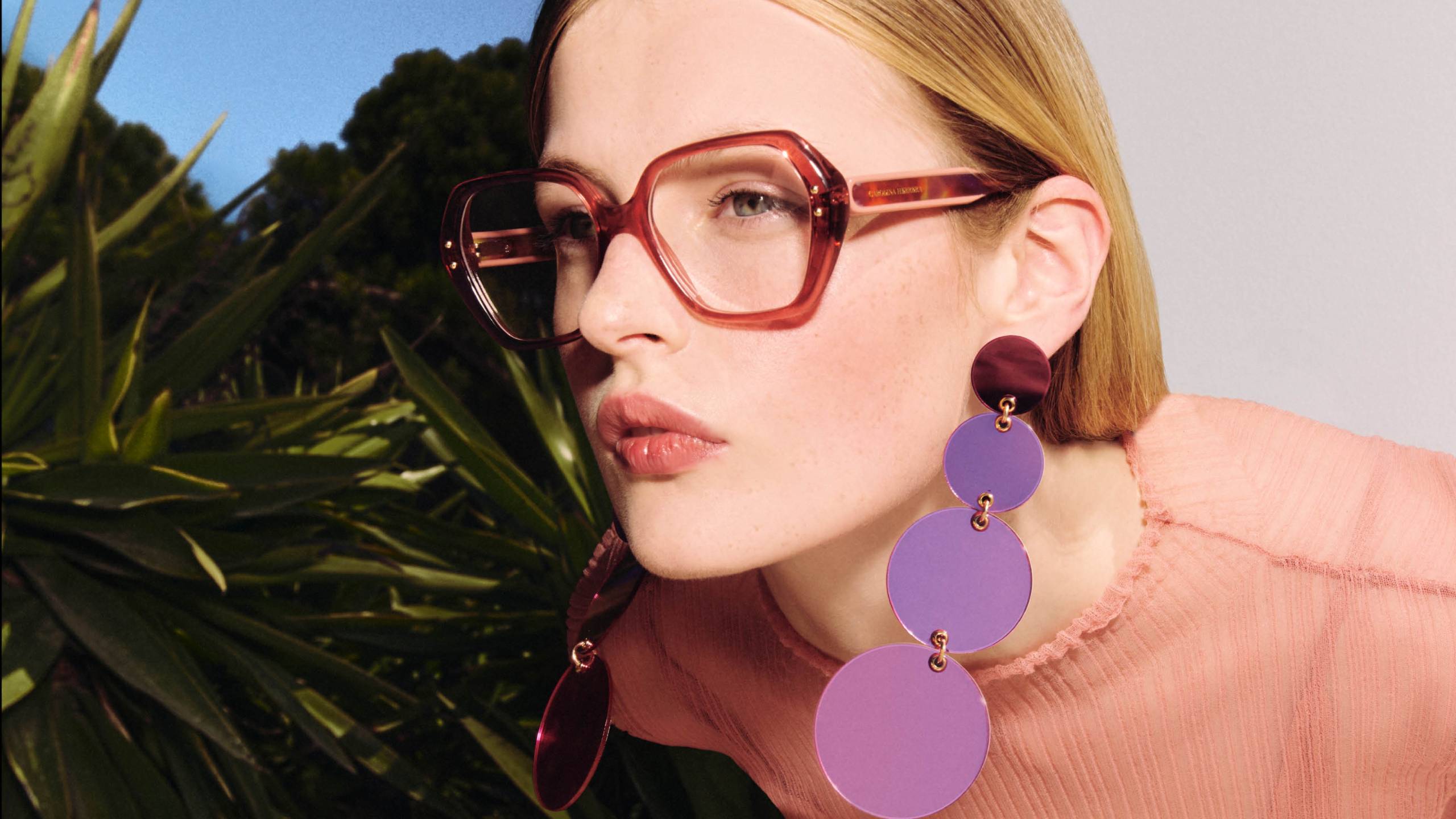Une femme blonde portant des lunettes à monture rouge surdimensionnée et des boucles d'oreilles circulaires violettes pose sur un fond de palmiers verts et de ciel bleu. Elle porte un pull en maille rose et affiche une expression subtile et confiante en regardant vers la camera