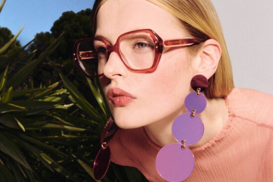 A blonde woman wearing oversized red-framed eyeglasses and statement purple circular drop earrings poses against a backdrop of green palm plants and blue sky. She's wearing a pink knit sweater and has a subtle, confident expression while looking toward the camera