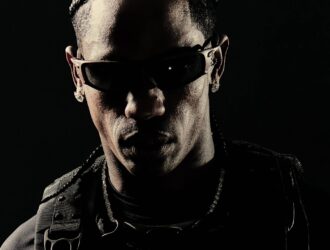 Main artcile image: Travis Scott wearing Oakley sunglasses in a dramatic black and white portrait, dressed in dark tactical-style clothing with straps and buckles against a black background, showcasing the post-apocalyptic aesthetic from his collaboration with Oakley