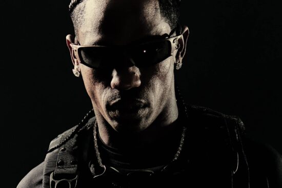 Main artcile image: Travis Scott wearing Oakley sunglasses in a dramatic black and white portrait, dressed in dark tactical-style clothing with straps and buckles against a black background, showcasing the post-apocalyptic aesthetic from his collaboration with Oakley