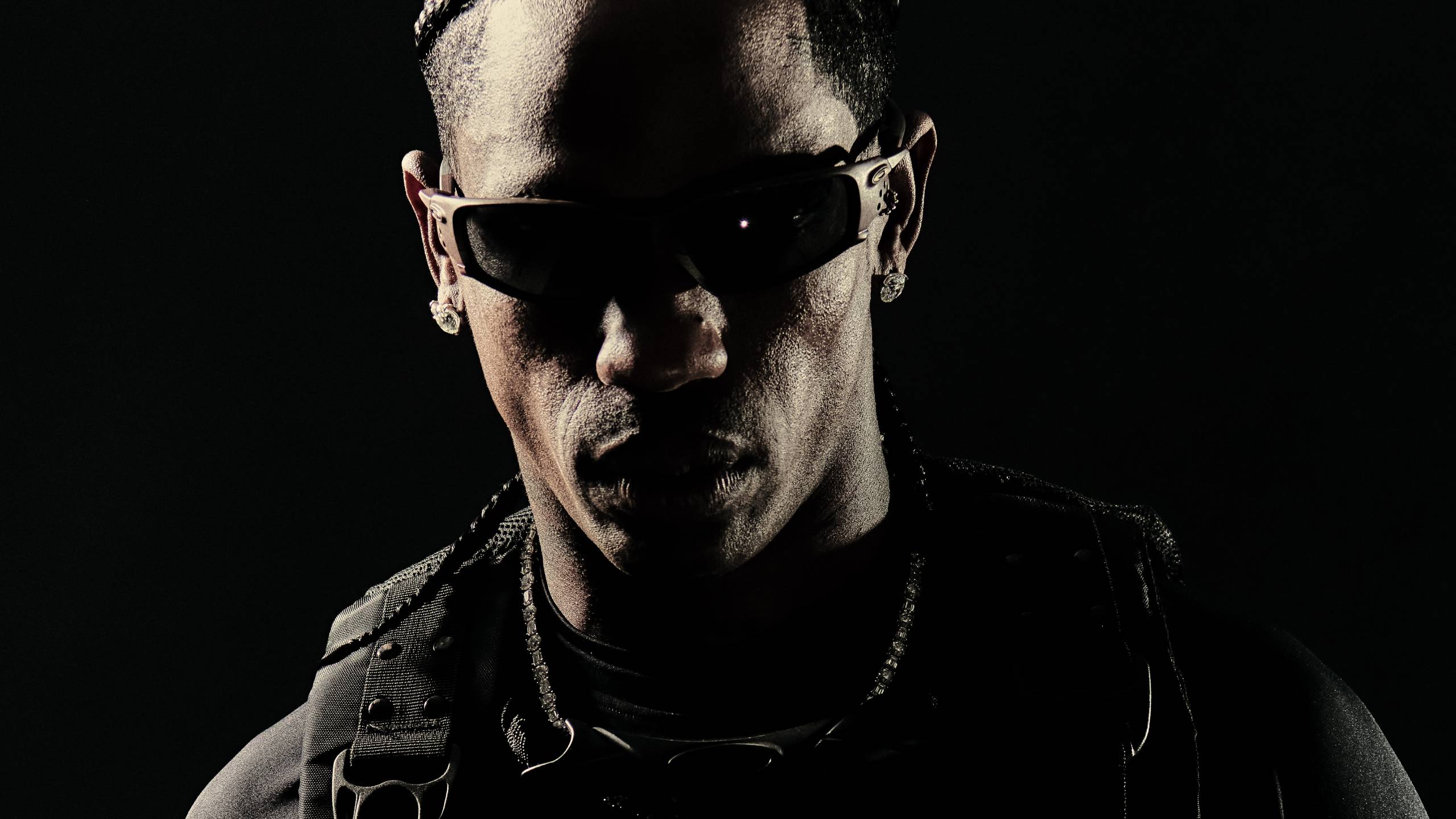 Main artcile image: Travis Scott wearing Oakley sunglasses in a dramatic black and white portrait, dressed in dark tactical-style clothing with straps and buckles against a black background, showcasing the post-apocalyptic aesthetic from his collaboration with Oakley