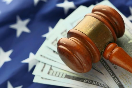 Image principale de l'article : Un marteau de juge en bois posé sur des billets de dollars américains avec un drapeau américain en arrière-plan, symbolisant l'intersection du droit et de la finance dans le système juridique américain.