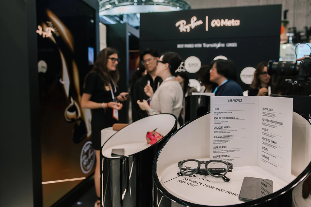 Stand d'exposition EssilorLuxottica au VivaTech 2025 avec des lunettes Ray-Ban Meta exposées sur des tables circulaires, menu petit-déjeuner visible, et visiteurs explorant le stand