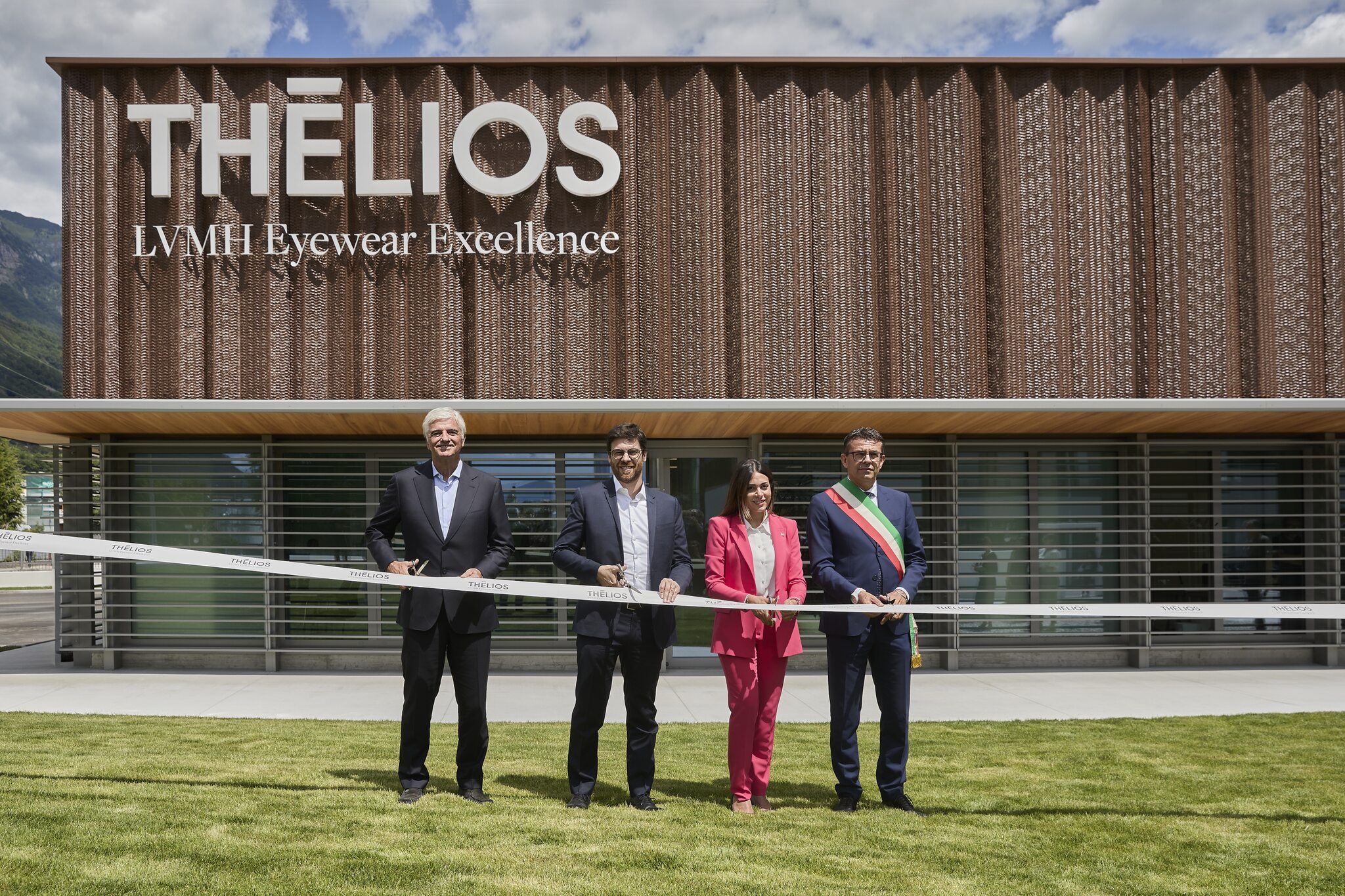 Here are the alt texts for both images in French and English: Image 1: English: "Ribbon-cutting ceremony at the new Thélios metal processing facility in Longarone, with four officials including Regional Councillor Valeria Mantovan in pink suit and local mayor with ceremonial sash standing in front of the modern building with wooden facade and mountain backdrop.