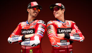 Two Ducati Lenovo Team MotoGP riders standing together with arms crossed, both wearing red and white racing leathers with sponsor logos and stylish sunglasses. They're smiling confidently against a deep red background, showcasing the CARRERA|DUCATI eyewear collection.