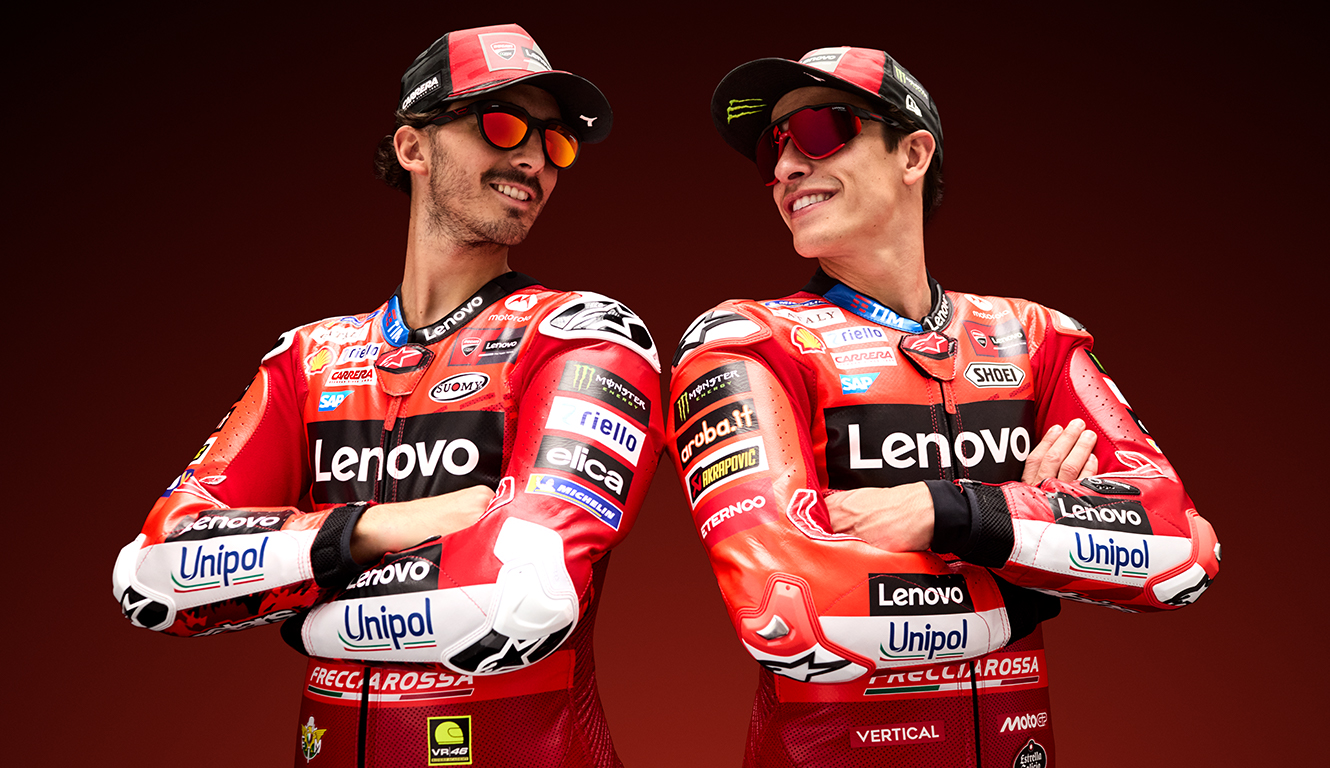 Deux pilotes MotoGP de l'équipe Ducati Lenovo debout ensemble, bras croisés, portant tous deux des combinaisons de course rouges et blanches avec logos de sponsors et des lunettes de soleil stylées. Ils sourient avec confiance sur un fond rouge profond, présentant la collection de lunettes CARRERA|DUCATI.