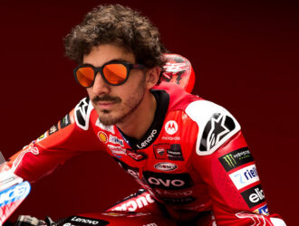 Gros plan d'un pilote MotoGP Ducati aux cheveux bouclés et à la barbe, portant des lunettes de soleil aux verres orangés et une combinaison de course rouge Ducati avec divers logos de sponsors incluant Lenovo, Shell et Monster Energy. Il est positionné sur sa moto sur un fond rouge.
