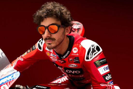 Gros plan d'un pilote MotoGP Ducati aux cheveux bouclés et à la barbe, portant des lunettes de soleil aux verres orangés et une combinaison de course rouge Ducati avec divers logos de sponsors incluant Lenovo, Shell et Monster Energy. Il est positionné sur sa moto sur un fond rouge.