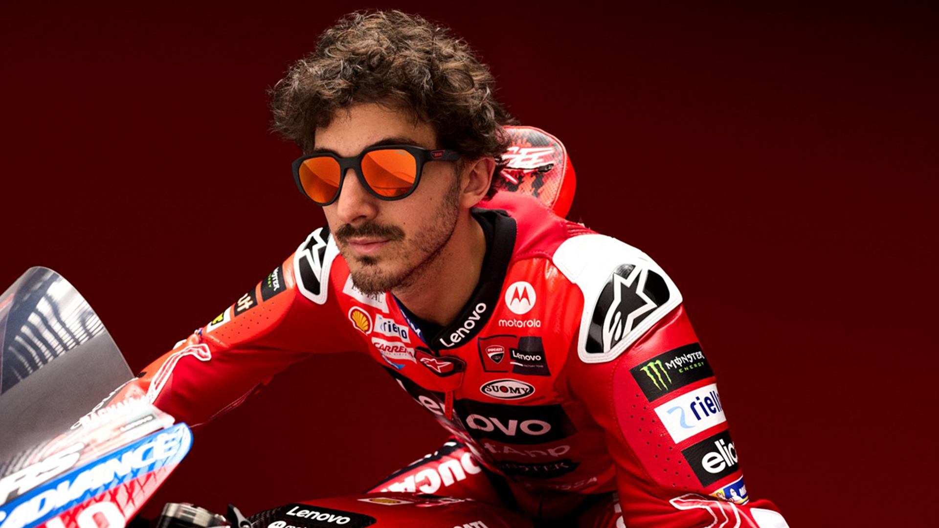 Gros plan d'un pilote MotoGP Ducati aux cheveux bouclés et à la barbe, portant des lunettes de soleil aux verres orangés et une combinaison de course rouge Ducati avec divers logos de sponsors incluant Lenovo, Shell et Monster Energy. Il est positionné sur sa moto sur un fond rouge.