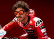 Close-up of a Ducati MotoGP rider with curly hair and facial hair, wearing orange-tinted sunglasses and red Ducati racing leathers with various sponsor logos including Lenovo, Shell, and Monster Energy. He's positioned on his motorcycle against a red background.