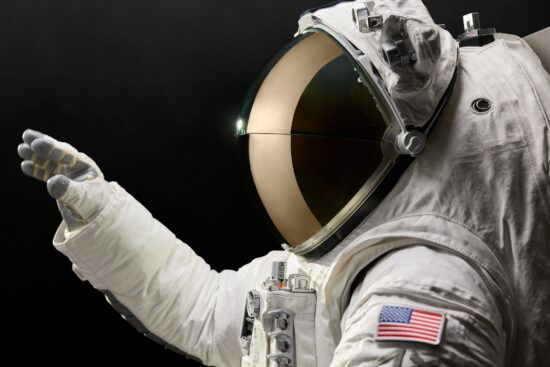 Astronaute en combinaison spatiale blanche avec casque à visière dorée sur fond noir, portant des gants et arborant un écusson du drapeau américain sur la manche, représentant la combinaison spatiale AxEMU d'Axiom Space avec la technologie de visière Oakley pour les missions lunaires.