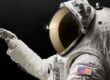 Astronaut in white spacesuit with gold-tinted visor helmet against black background, wearing gloves and displaying American flag patch on sleeve, representing the Axiom Space AxEMU spacesuit with Oakley visor technology for lunar missions.