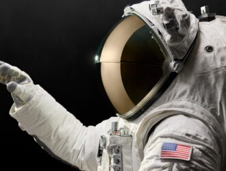 Astronaut in white spacesuit with gold-tinted visor helmet against black background, wearing gloves and displaying American flag patch on sleeve, representing the Axiom Space AxEMU spacesuit with Oakley visor technology for lunar missions.