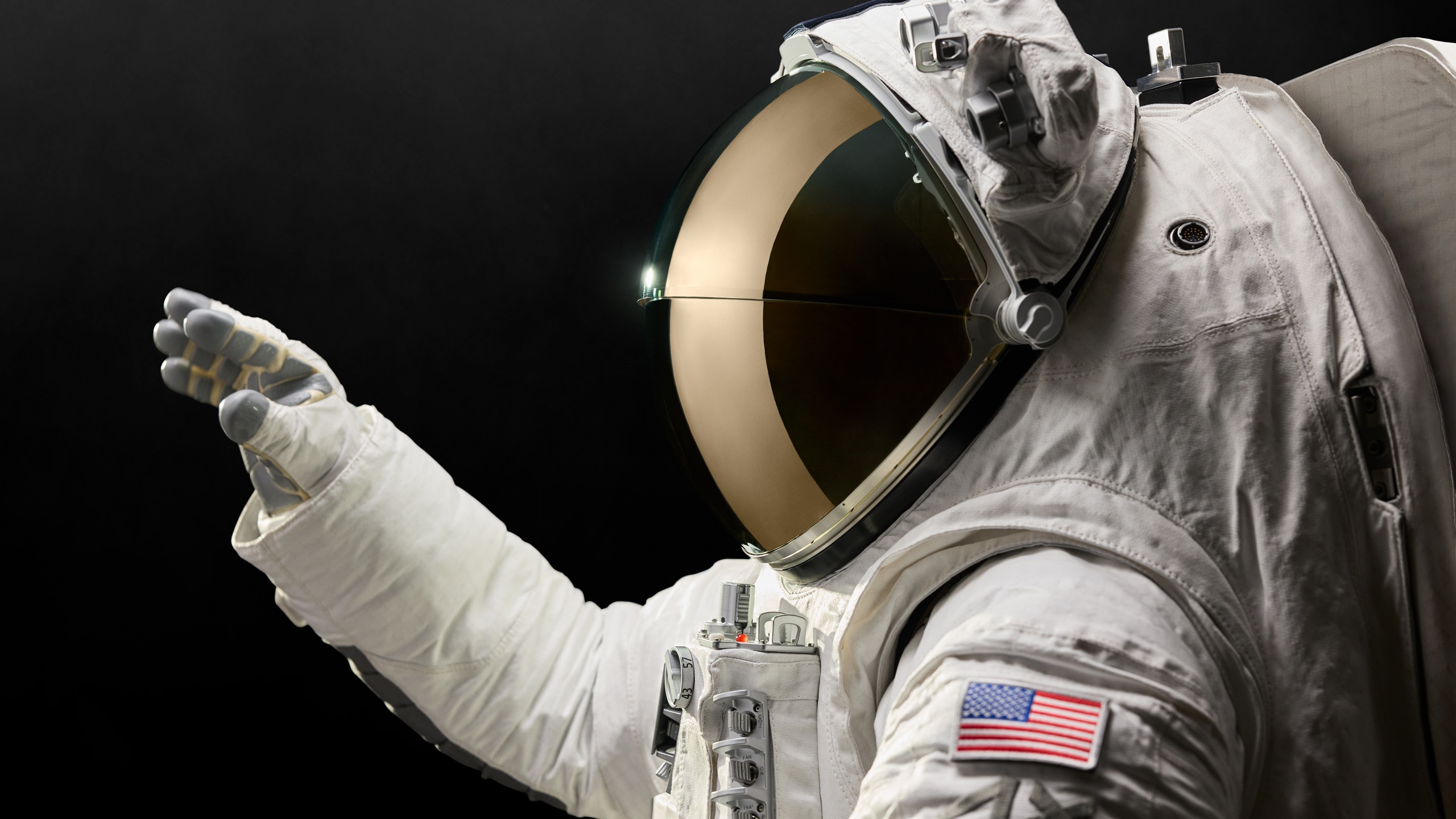 Astronaut in white spacesuit with gold-tinted visor helmet against black background, wearing gloves and displaying American flag patch on sleeve, representing the Axiom Space AxEMU spacesuit with Oakley visor technology for lunar missions.
