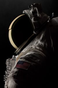 Vue de profil rapprochée d'un astronaute en combinaison spatiale avec visière de casque teintée dorée sur fond noir, montrant les mécanismes détaillés du casque et l'écusson du drapeau américain sur la manche, mettant en évidence la technologie de visière Oakley pour la combinaison spatiale AxEMU.
