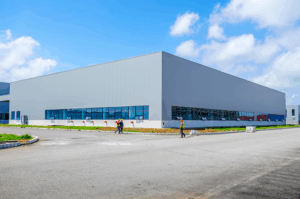 Modern industrial warehouse facility with gray exterior walls and large blue-tinted windows, featuring workers in safety vests walking along the building's perimeter under a partly cloudy sky.