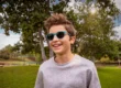 A young boy with tousled brown hair wearing blue sunglasses and a light gray sweatshirt smiles brightly at the camera. He's standing outdoors in a park setting with green grass, leafy trees, and a bright sunny sky with white clouds in the background.