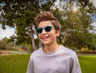 A young boy with tousled brown hair wearing blue sunglasses and a light gray sweatshirt smiles brightly at the camera. He's standing outdoors in a park setting with green grass, leafy trees, and a bright sunny sky with white clouds in the background.