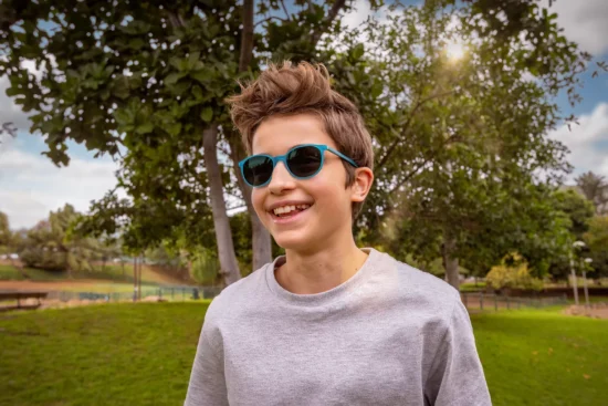 A young boy with tousled brown hair wearing blue sunglasses and a light gray sweatshirt smiles brightly at the camera. He's standing outdoors in a park setting with green grass, leafy trees, and a bright sunny sky with white clouds in the background.