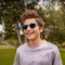 A young boy with tousled brown hair wearing blue sunglasses and a light gray sweatshirt smiles brightly at the camera. He's standing outdoors in a park setting with green grass, leafy trees, and a bright sunny sky with white clouds in the background.