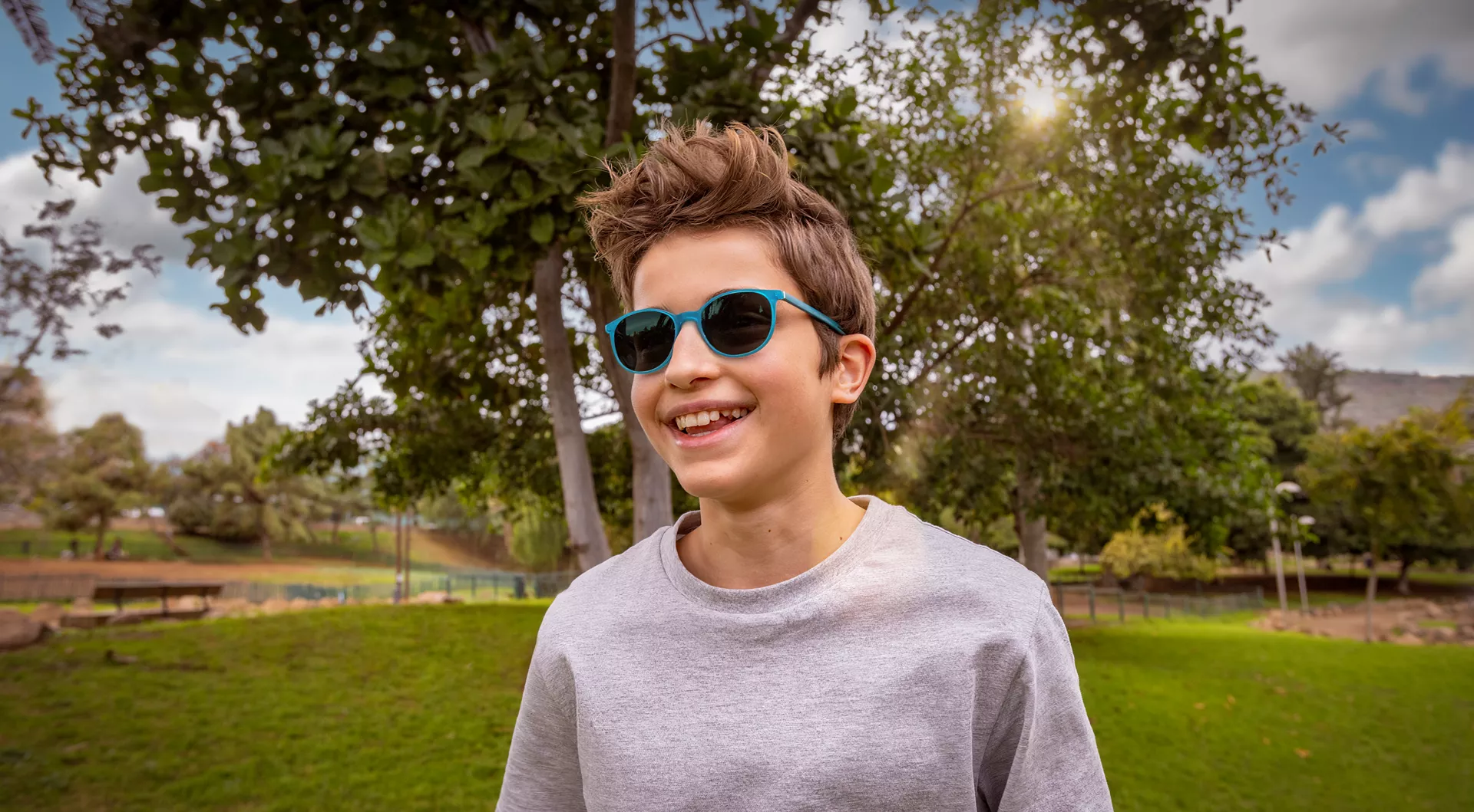 Un jeune garçon aux cheveux bruns ébouriffés portant des lunettes de soleil bleues et un sweat-shirt gris clair sourit largement à la caméra. Il se tient dehors dans un parc avec de l'herbe verte, des arbres feuillus, et un ciel ensoleillé lumineux avec des nuages blancs en arrière-plan.