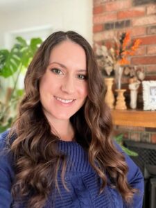 Kim Kochendorfer, responsable des affaires professionnelles chez HOYA Vision Canada, aux longs cheveux bruns ondulés, vêtue d'un pull bleu, sourit dans un décor intérieur chaleureux avec une cheminée en briques et une décoration sur le thème de l'automne en arrière-plan.