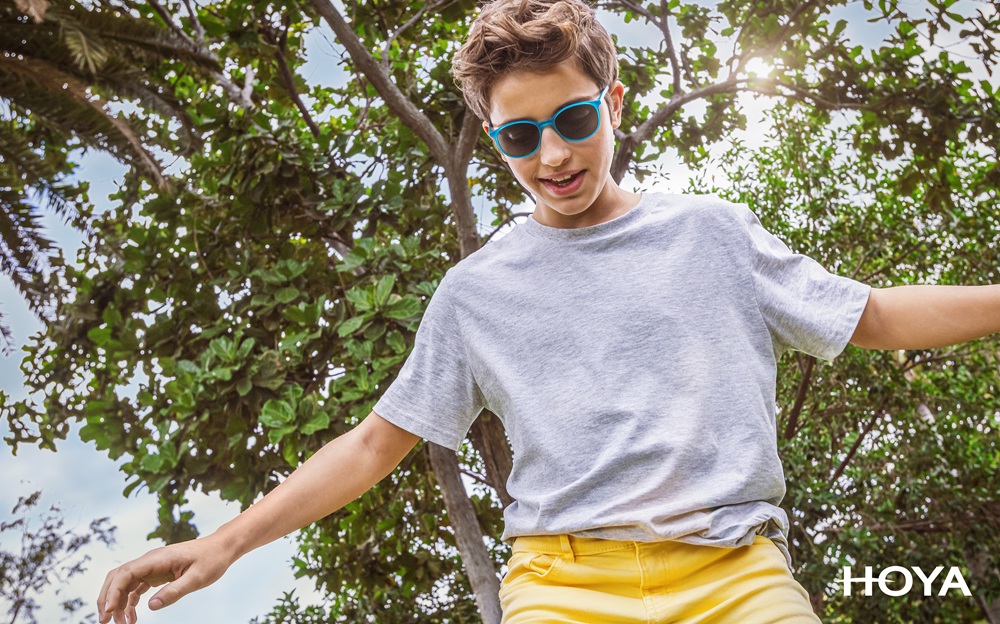 Boy wearing blue sunglasses, a gray t-shirt, and yellow shorts balancing playfully outdoors on a sunny day, with leafy green trees in the background. HOYA logo appears in the bottom right corner.