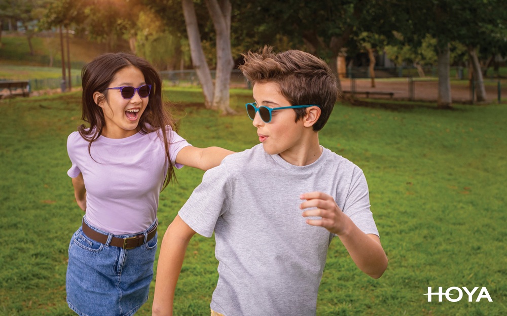 Two children wearing sunglasses playfully running in a sunny park, smiling and laughing; the girl wears a light purple shirt and denim skirt, and the boy wears a gray t-shirt. HOYA logo is visible in the corner.