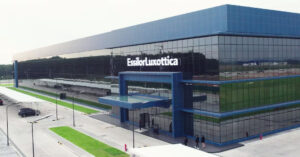Modern EssilorLuxottica corporate facility with contemporary glass and steel architecture featuring blue and green accents, large windows, and the company name prominently displayed on the building exterior.