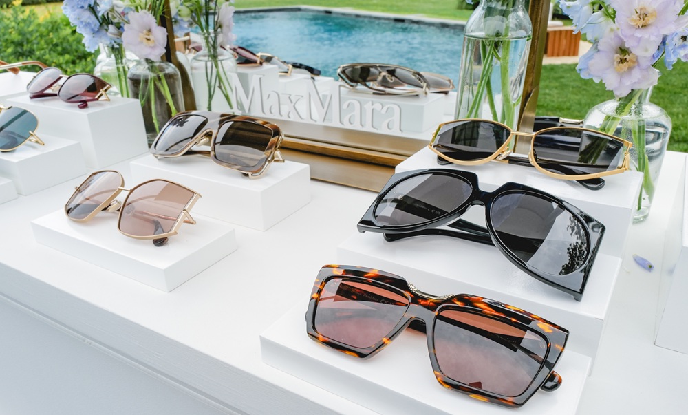 Display of Max Mara SS25 sunglasses collection featuring various styles including oversized frames, cat-eye shapes, and geometric designs in different colors and finishes, arranged on white display stands with floral arrangements and pool setting in background.
