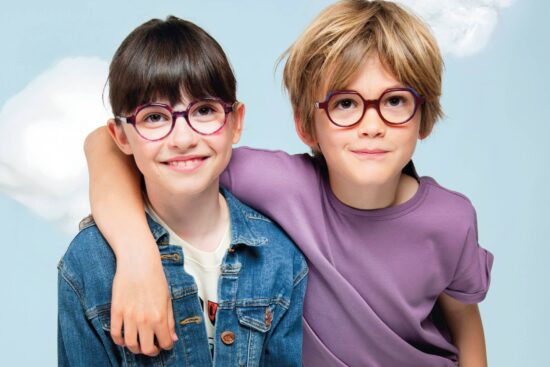 Deux enfants souriants portant des lunettes colorées J.F. Rey - une fille en veste en jean avec des montures rondes rose et bleue, et un garçon en t-shirt violet avec des montures rondes bordeaux, posant ensemble devant un arrière-plan de ciel nuageux