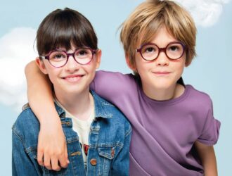 Two smiling children wearing colorful J.F. Rey eyeglasses - a girl in a denim jacket with round pink and blue frames, and a boy in a purple shirt with round burgundy frames, posed together against a soft cloudy sky background