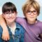 Two smiling children wearing colorful J.F. Rey eyeglasses - a girl in a denim jacket with round pink and blue frames, and a boy in a purple shirt with round burgundy frames, posed together against a soft cloudy sky background