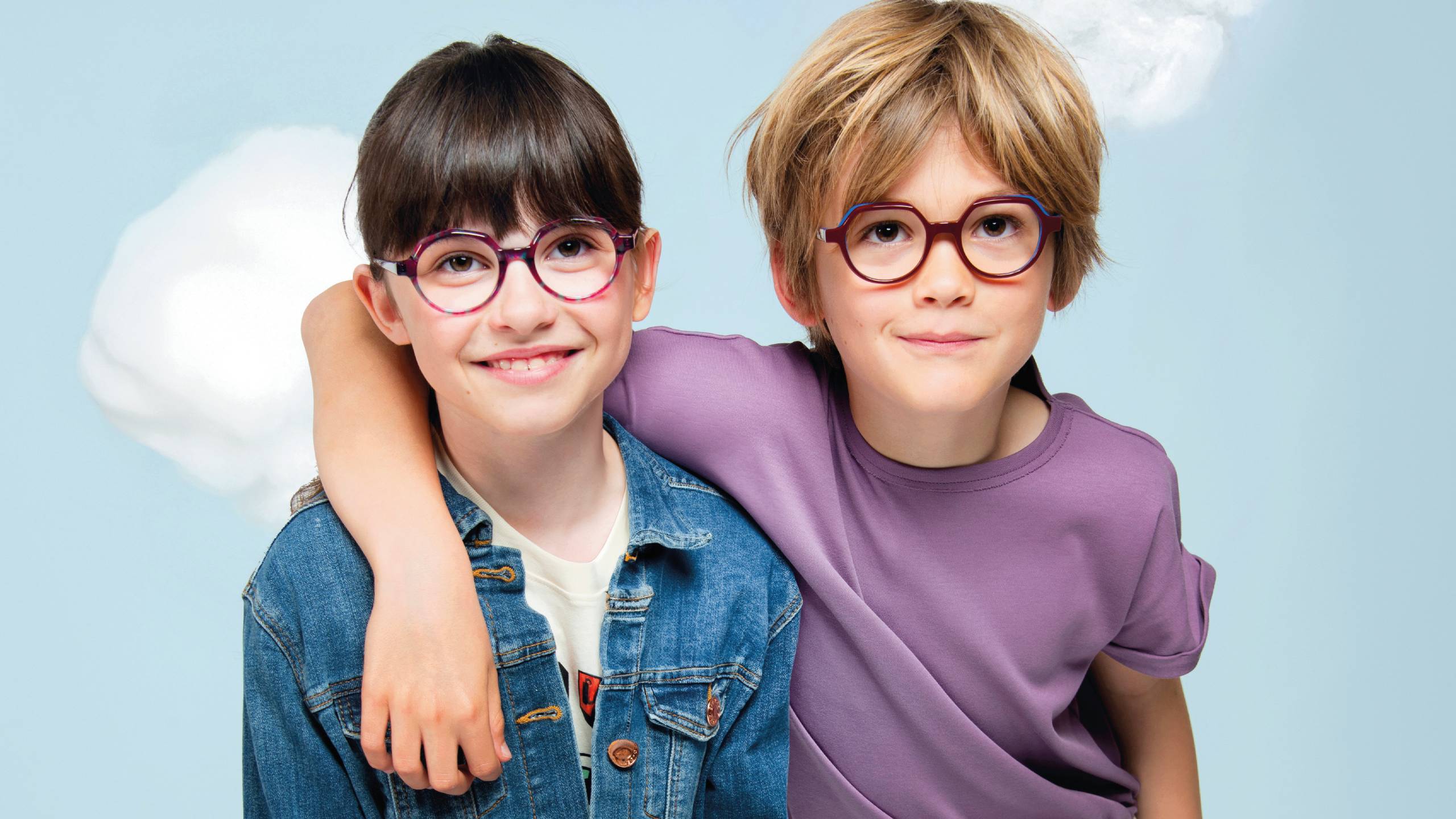 Two smiling children wearing colorful J.F. Rey eyeglasses - a girl in a denim jacket with round pink and blue frames, and a boy in a purple shirt with round burgundy frames, posed together against a soft cloudy sky background