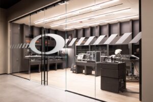 Glass-enclosed Oakley performance optics laboratory and manufacturing space with industrial equipment, testing stations, and the iconic Oakley 'O' logo prominently displayed.