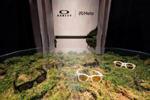 Oakley Meta smart glasses display featuring white-framed eyewear positioned on a moss-covered landscape diorama with natural elements and Oakley/Meta partnership branding.