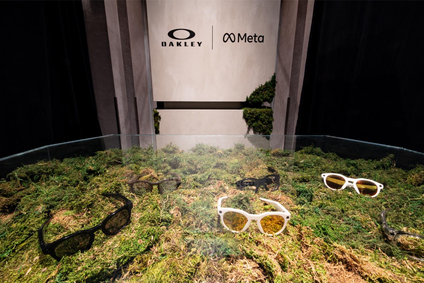 Oakley Meta smart glasses display featuring white-framed eyewear positioned on a moss-covered landscape diorama with natural elements and Oakley/Meta partnership branding.