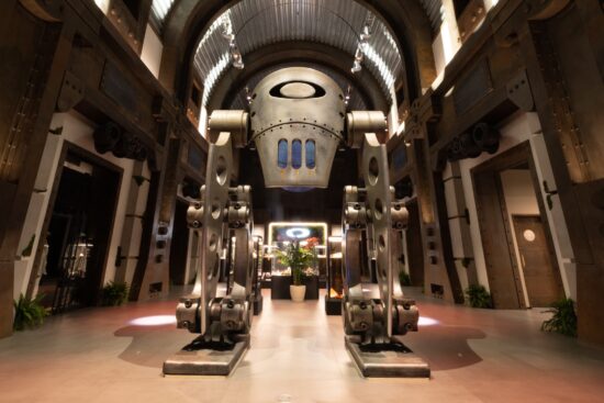 Entrée futuriste du siège social d'Oakley avec une grande sculpture métallique ressemblant à un robot arborant le logo Oakley, des éléments de design industriel et un éclairage dramatique dans un cadre d'atrium.