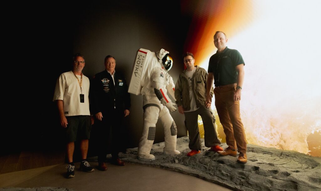 Quatre hommes posant avec une combinaison spatiale d'Axiom Space exposée dans ce qui semble être un centre spatial ou un musée, avec un terrain lunaire simulé et un éclairage atmosphérique en arrière-plan, mettant en valeur le partenariat entre Oakley et Axiom Space.