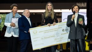 Close-up view of the Sergio Cereda Eyewear Design Award ceremony with officials presenting the $10,000 first place winner's check dated February 8, 2025. The award winner, a young woman in black lace attire, holds the ceremonial check alongside award presenters at the MIDO exhibition.