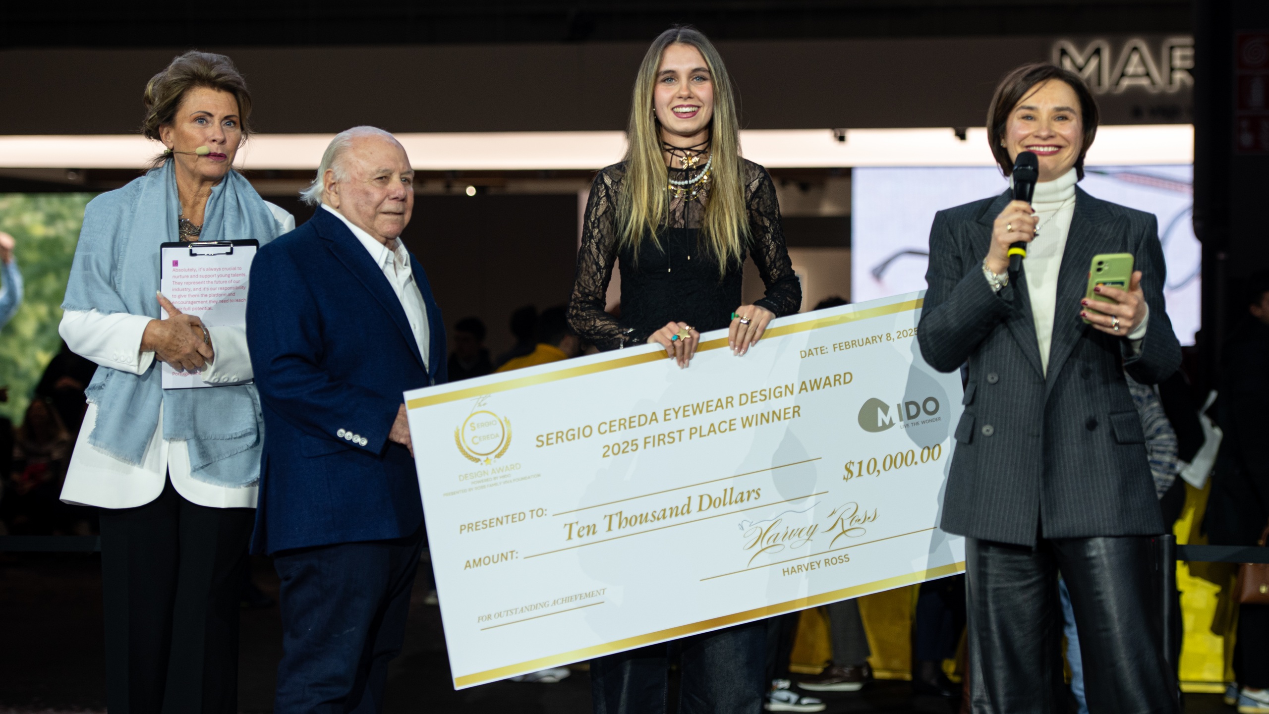 Close-up view of the Sergio Cereda Eyewear Design Award ceremony with officials presenting the $10,000 first place winner's check dated February 8, 2025. The award winner, a young woman in black lace attire, holds the ceremonial check alongside award presenters at the MIDO exhibition.