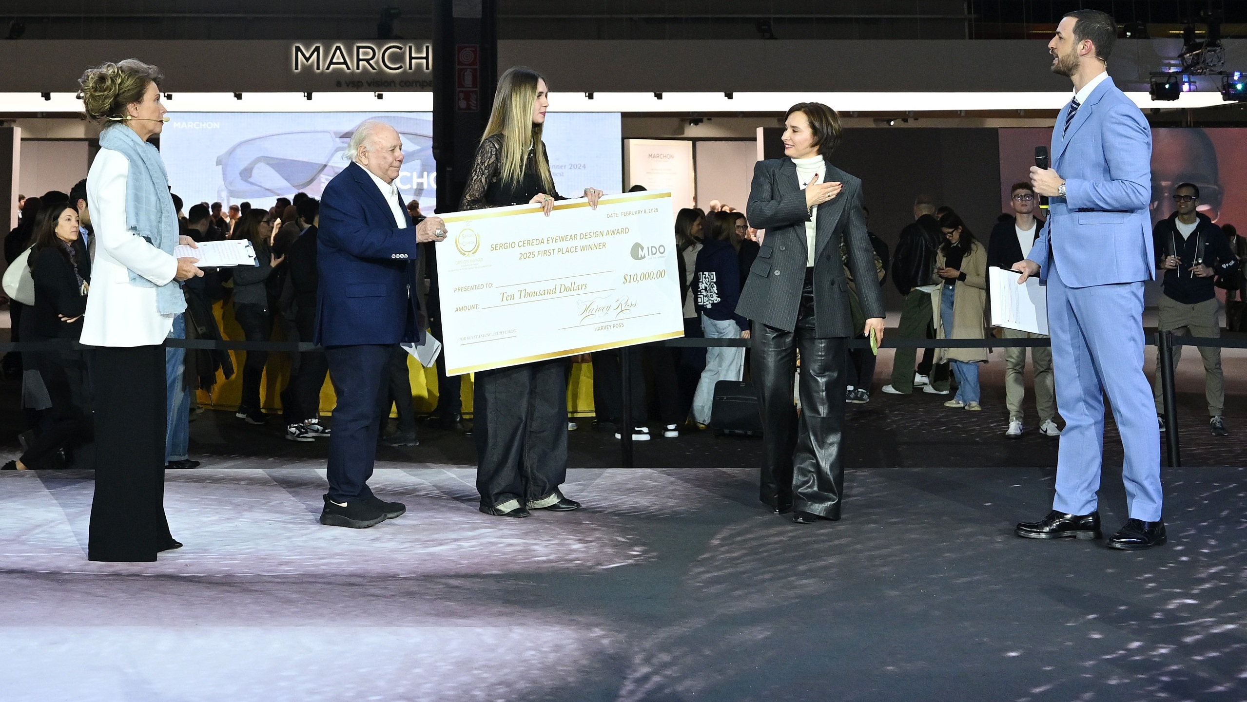 Award ceremony at MIDO 2025 showing the presentation of the Sergio Cereda Eyewear Design Award. A large ceremonial check for $10,000 is being presented to the first place winner, with several officials in formal attire participating in the presentation on stage.