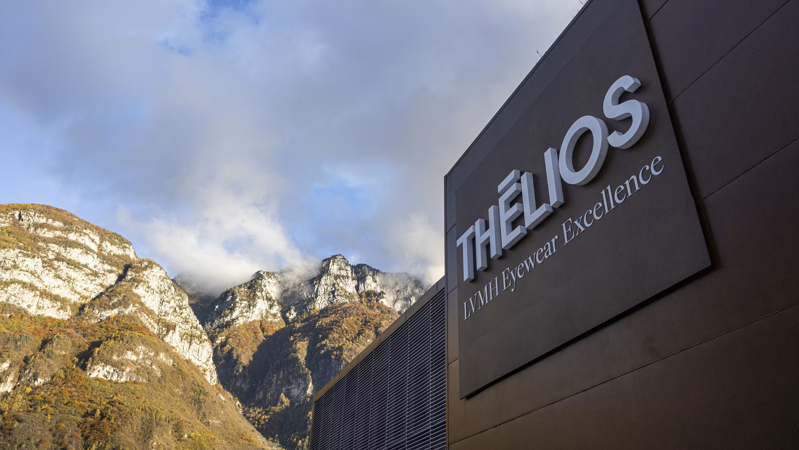 Vue extérieure de l'installation Thélios LVMH Eyewear Excellence à Longarone, montrant le bâtiment moderne sombre avec la signalétique de l'entreprise sur le fond dramatique des montagnes des Dolomites italiennes.
