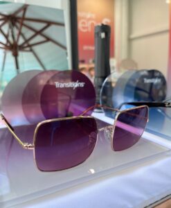 Close-up of elegant gold-framed sunglasses with purple gradient Transitions lenses displayed on a clear surface, with Transitions branded lenses visible in the blurred background.