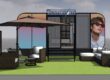 3D rendering of a modern Transitions outdoor exhibition booth featuring a green carpet area with white seating, umbrellas, and large displays showing a person wearing Transitions lenses, set against a cloudy sky backdrop.